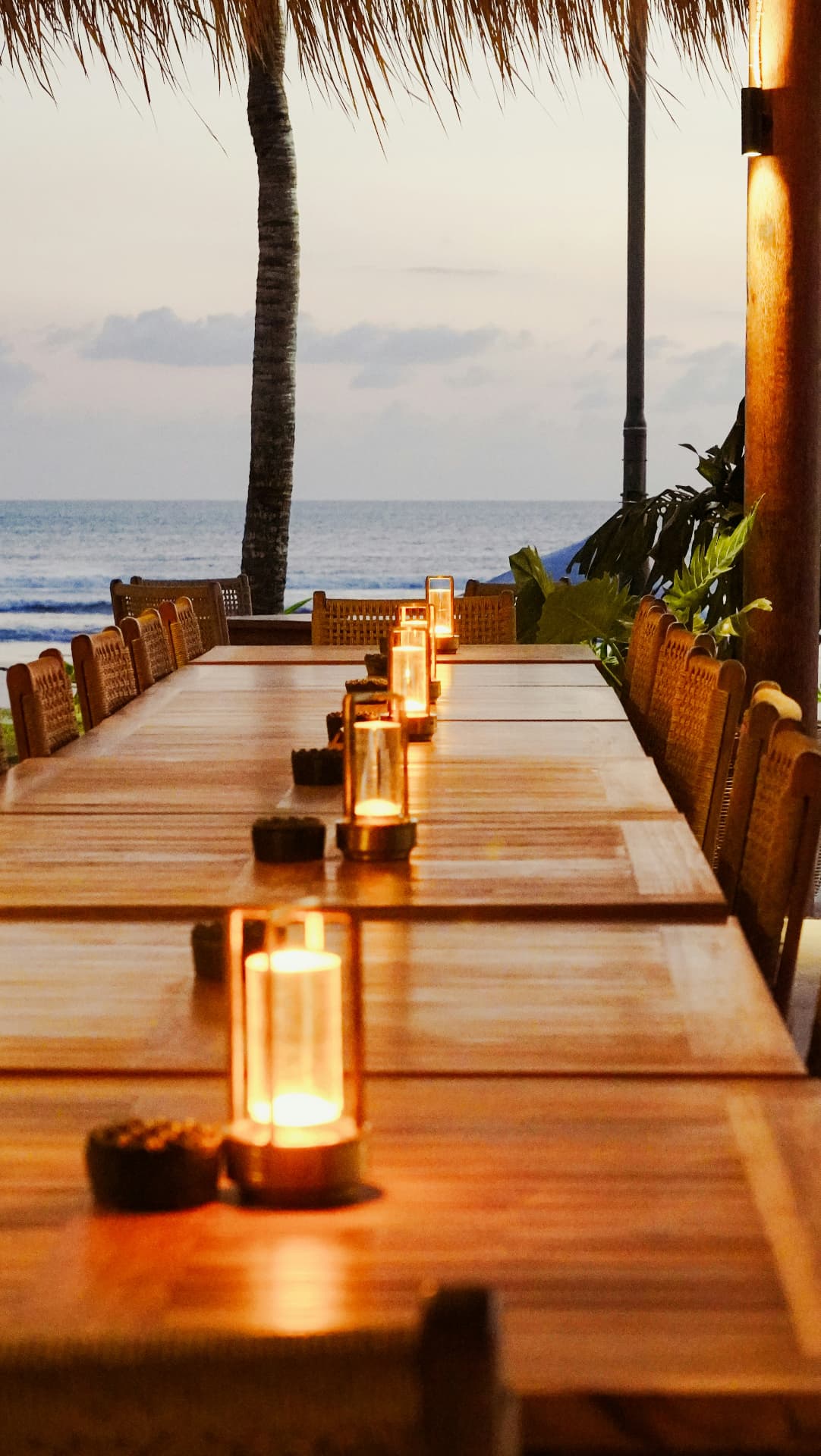 An empty long candlelit table beneath palm trees, with the ocean beyond